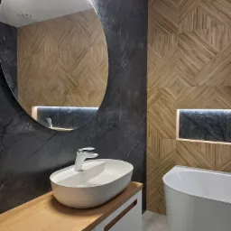 Nowoczesna łazienka z owalną umywalką na drewnianym blacie, okrągłym lustrem z podświetleniem LED, czarnymi płytkami imitującymi marmur i drewnopodobnymi płytkami ułożonymi w geometryczny wzór...