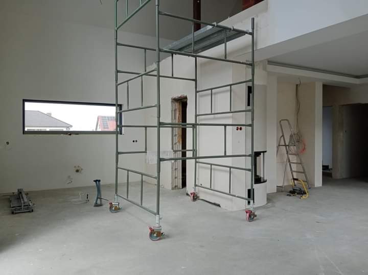 Wnętrze w trakcie remontu z metalowym rusztowaniem na kółkach, widoczne narzędzia i drabina oparta o ścianę, okno z widokiem na dom.