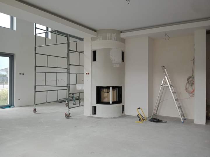 Wnętrze w trakcie prac wykończeniowych: okrągły kominek z czarnym wkładem, rusztowanie na kółkach, drabina aluminiowa, surowe ściany przygotowane pod malowanie, widoczne kable elektryczne.