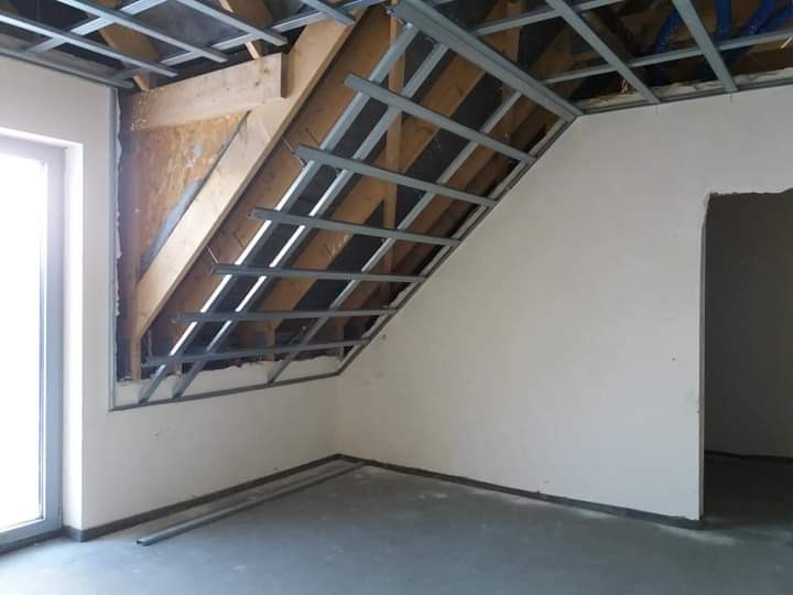 Montaż metalowej konstrukcji pod płytę gipsowo-kartonową na poddaszu, widoczne drewniane elementy konstrukcyjne dachu i surowy stan ścian, przygotowanie do dalszych prac wykończeniowych.