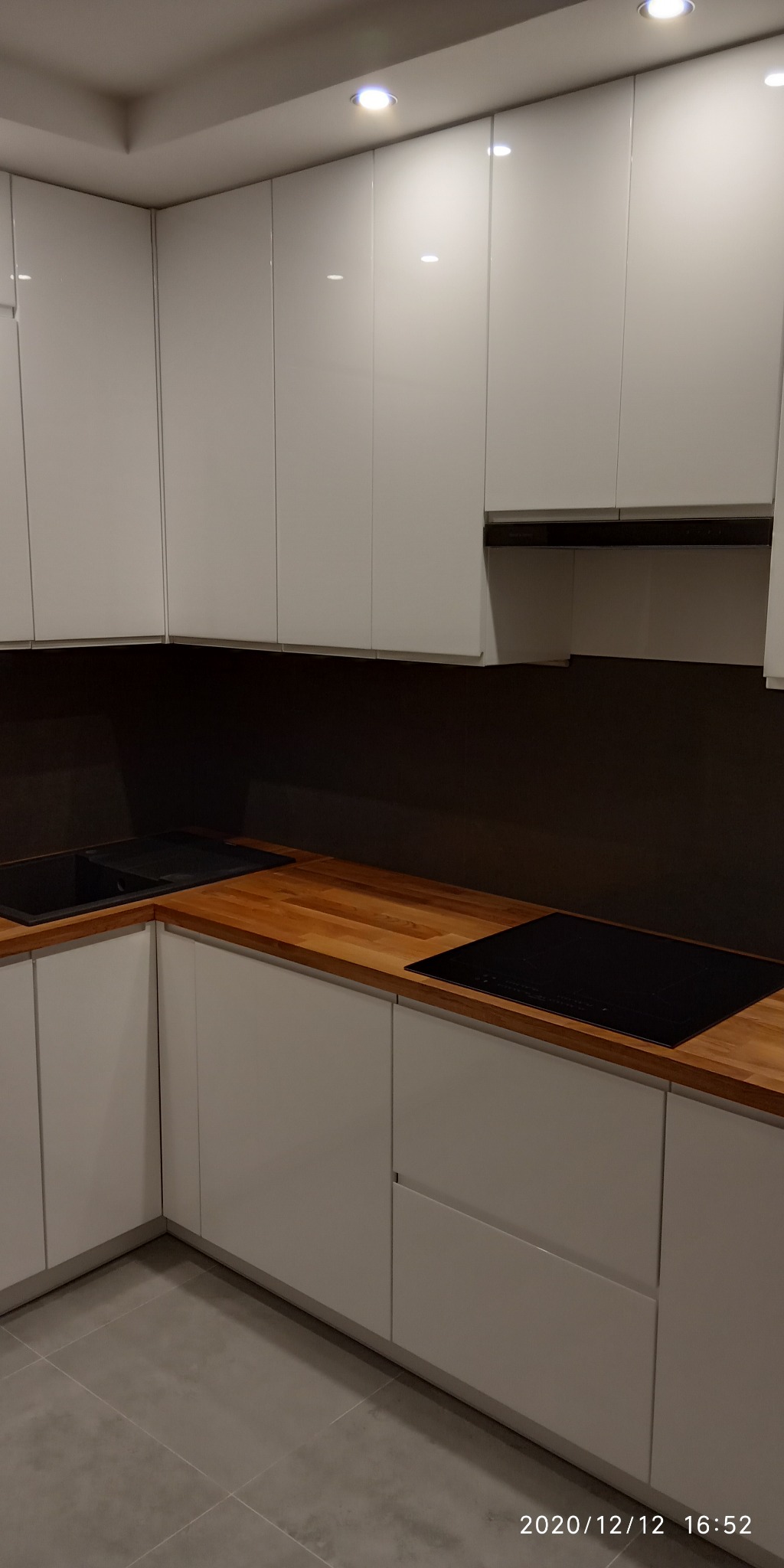 Nowoczesna kuchnia z białymi, lakierowanymi frontami szafek, drewnianym blatem i czarną płytą indukcyjną, oświetlona punktowymi lampami sufitowymi.