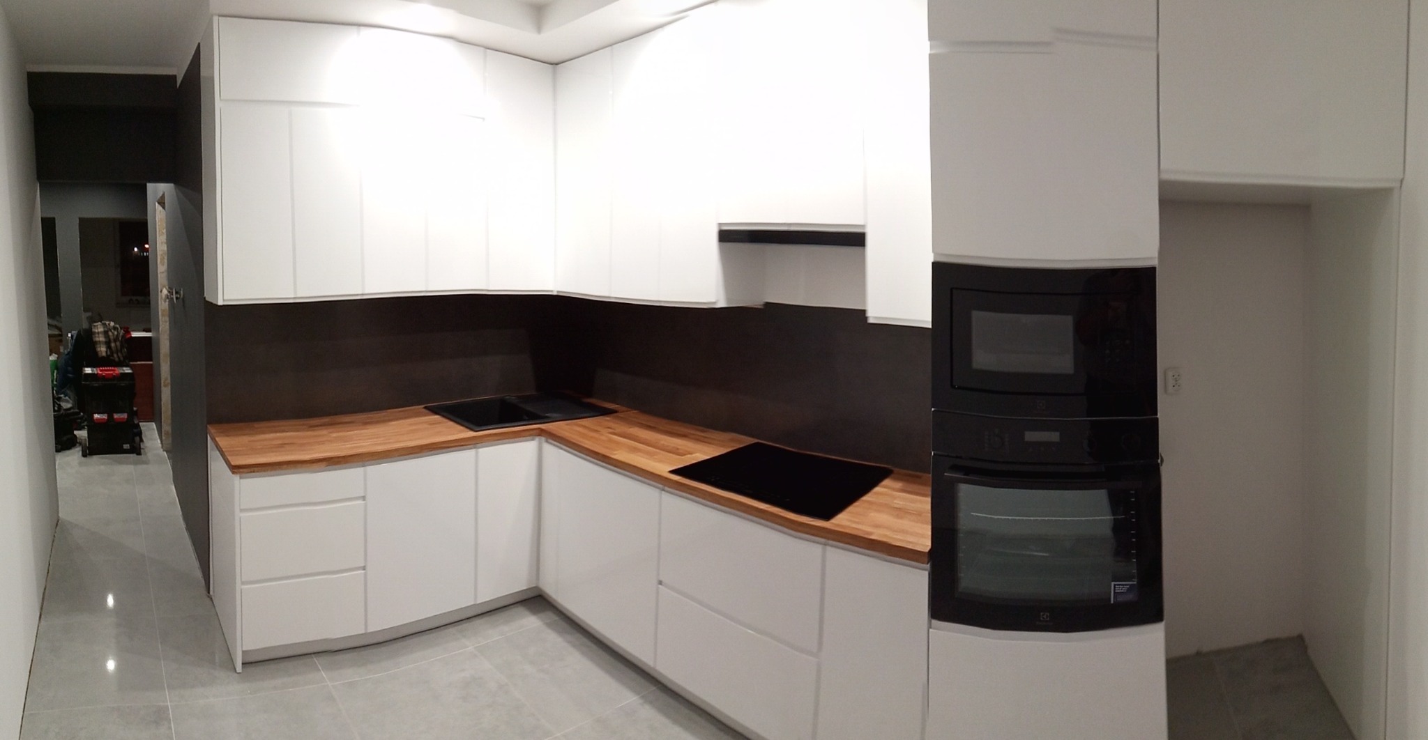 Nowoczesna kuchnia z białymi frontami i drewnianym blatem, czarne AGD w zabudowie, szara ściana nad blatem, widok na korytarz z narzędziami w tle.