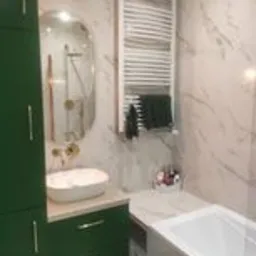 Nowoczesna łazienka w Środzie Śląskiej z białymi marmurowymi płytkami, zielonymi szafkami, owalnym lustrem i grzejnikiem drabinkowym. Umywalka nablatowa w kolorze ecru.