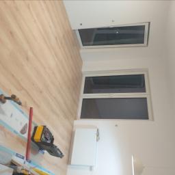 ANDRE - MONT Andrzej Strembicki - Wnętrze pokoju z dwoma oknami, drewnianą podłogą i narzędziami do montażu okien rozłożonymi na podłodze, w tym wiertarka i skrzynka narzędziowa.