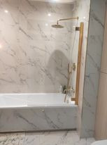 Biała wanna obłożona marmurowymi płytkami z złotą armaturą i szklanym parawanem.