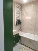 Nowoczesna łazienka z zielonymi szafkami, marmurowymi płytkami na ścianach i zabudowaną wanną. Biały grzejnik drabinkowy z czarnymi ręcznikami. Umywalka nablatowa z beżowym blatem.