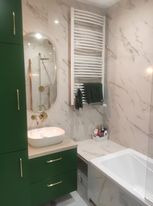 Nowoczesna łazienka w Środzie Śląskiej z białymi marmurowymi płytkami, zielonymi szafkami, owalnym lustrem i grzejnikiem drabinkowym. Umywalka nablatowa w kolorze ecru.