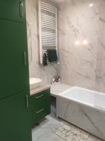 Wykończona łazienka z białymi marmurkowymi płytkami, zielonymi szafkami, białą umywalką nablatową i grzejnikiem drabinkowym z ręcznikami. Widoczna wanna obudowana płytkami w tym samym stylu.
