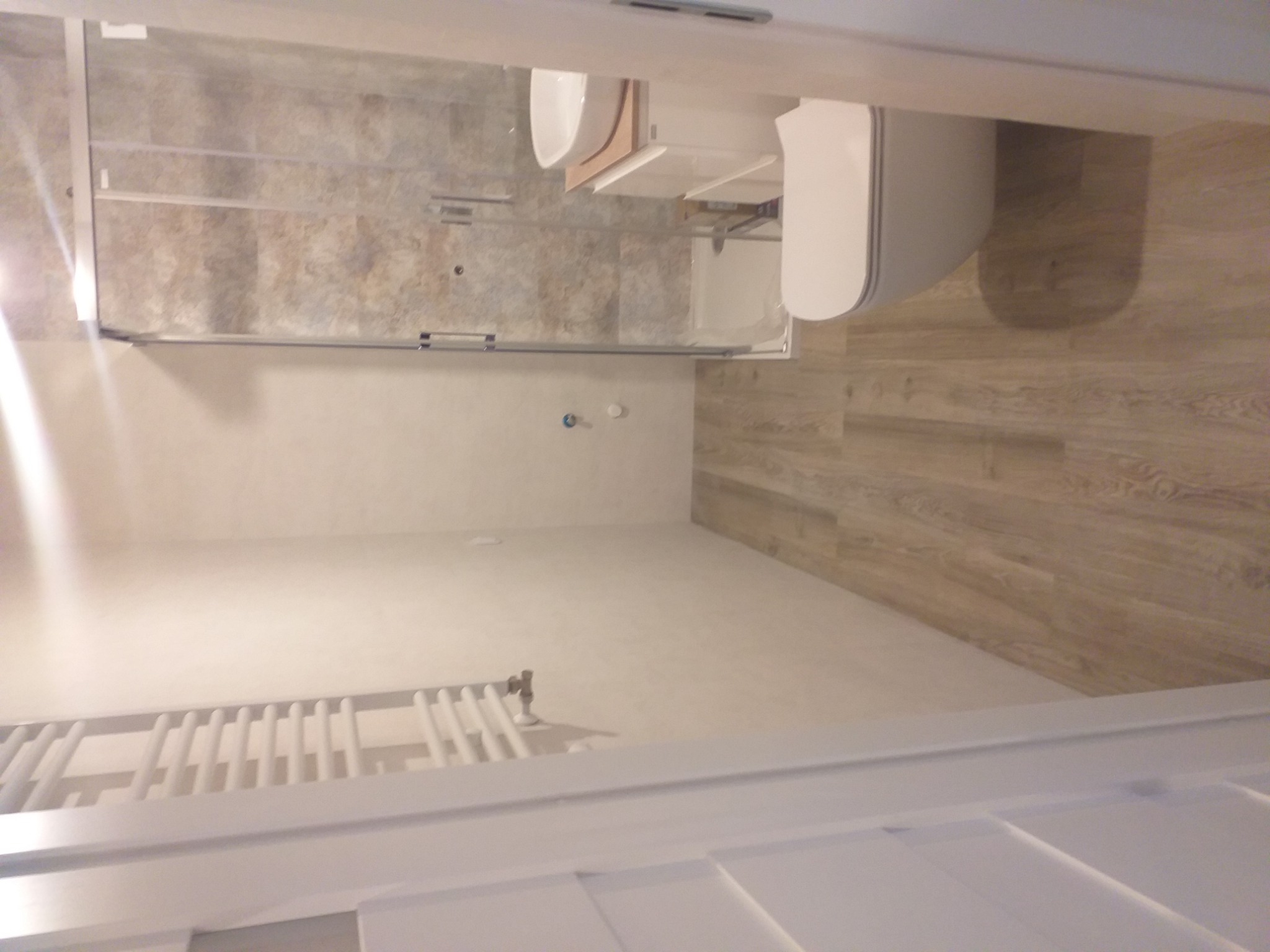 Ukończona łazienka z kabiną prysznicową z przesuwnymi drzwiami, wiszącą toaletą, grzejnikiem drabinkowym i podłogą imitującą drewno, widok zza progu.