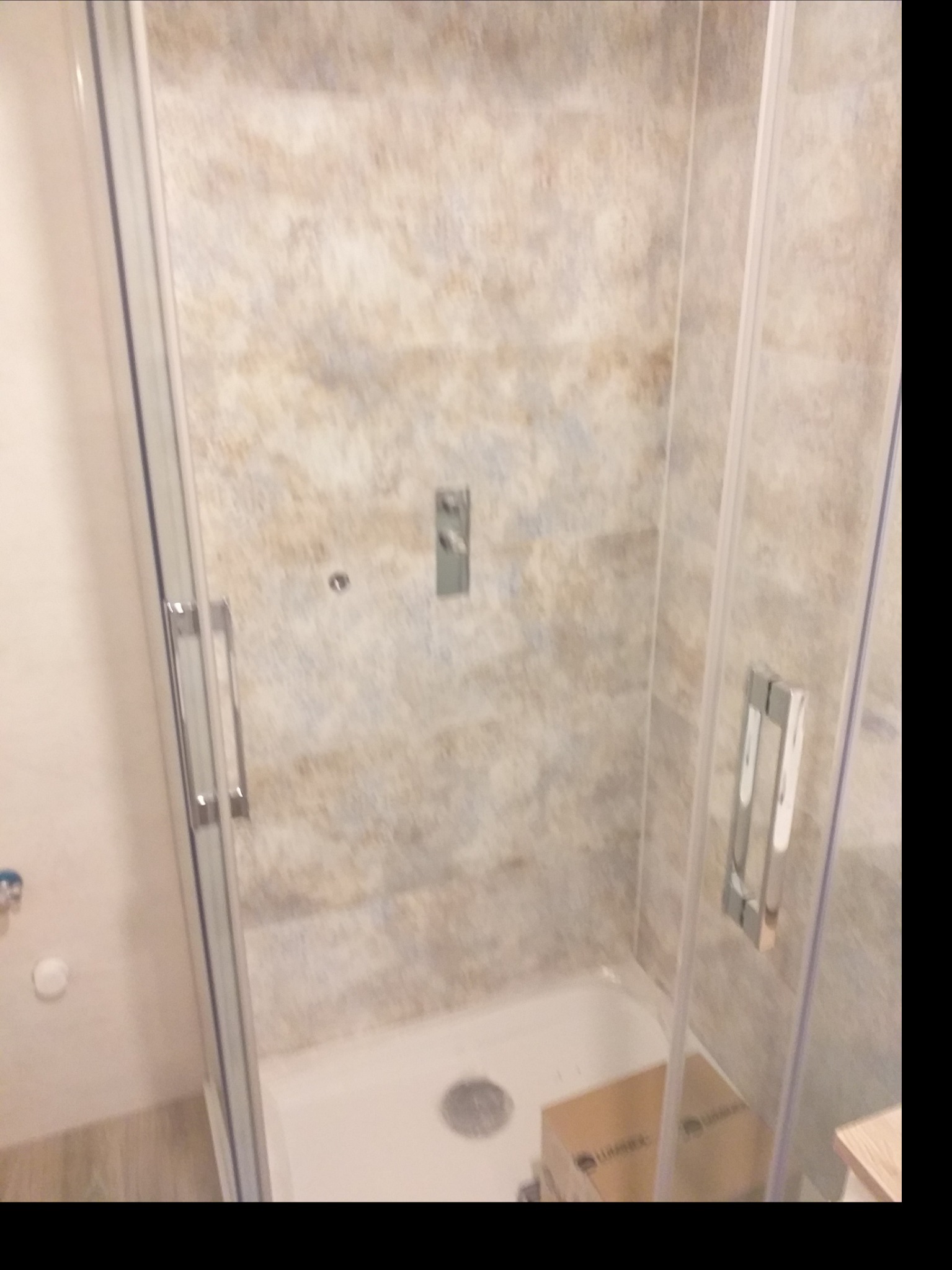 Wykończona kabina prysznicowa z beżowymi płytkami imitującymi kamień, widoczny chromowany panel sterowania i srebrne uchwyty, z pudełkiem na dnie brodzika.