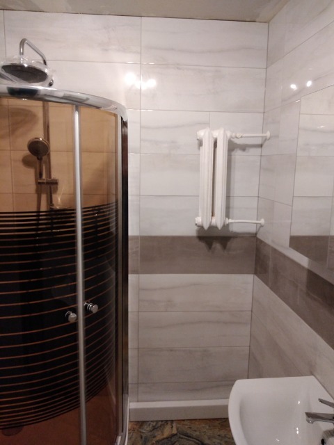 Ukończona łazienka z kabiną prysznicową z czarnymi paskami, grzejnikiem żeberkowym i umywalką, wykończona płytkami w odcieniach bieli i szarości z poziomym pasem dekoracyjnym.