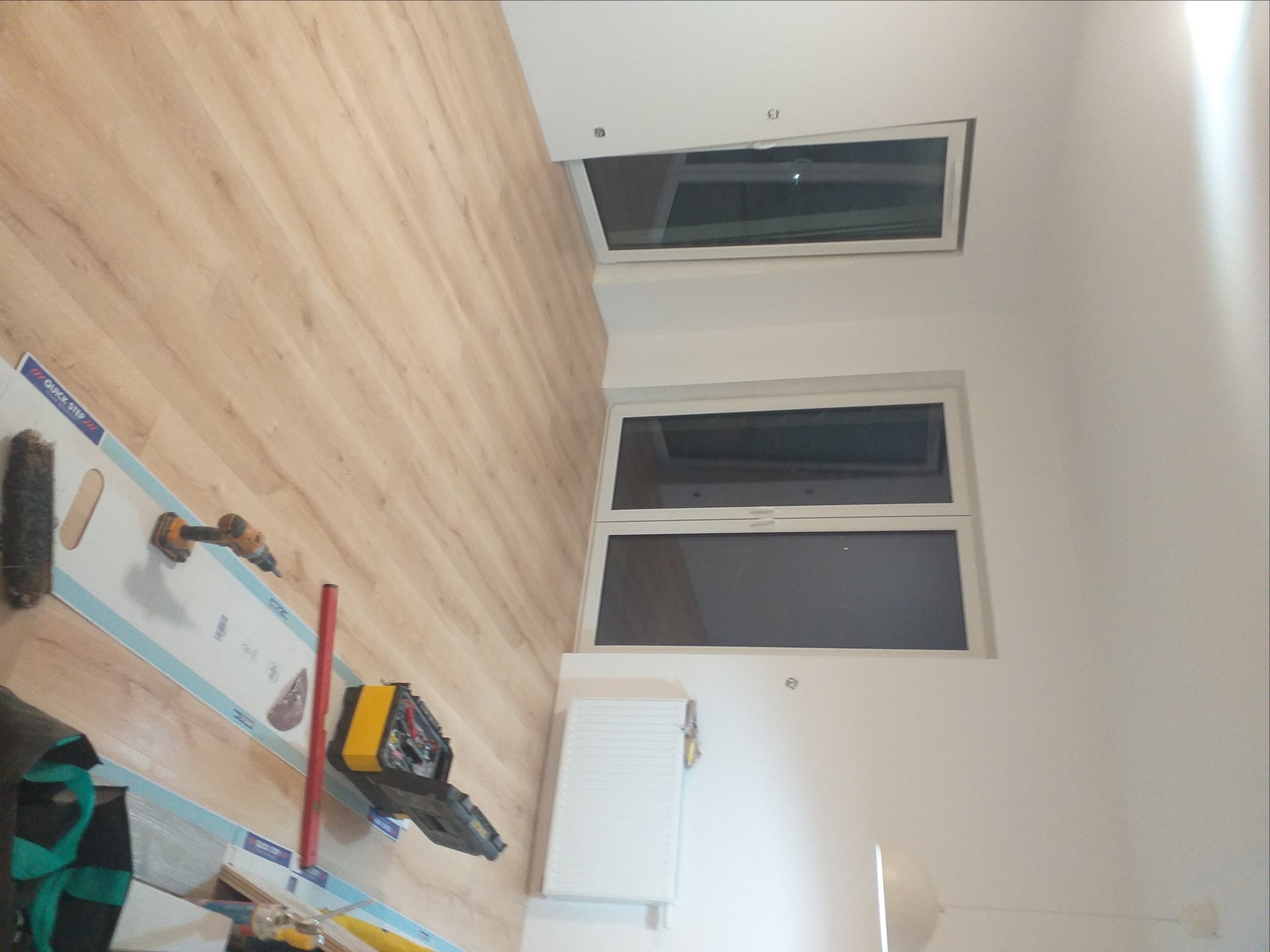 Wnętrze pokoju z dwoma oknami, drewnianą podłogą i narzędziami do montażu okien rozłożonymi na podłodze, w tym wiertarka i skrzynka narzędziowa.