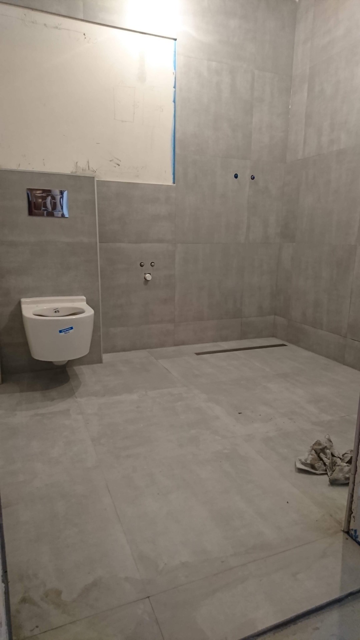 Szara łazienka w trakcie remontu, widoczna umywalka podwieszana, odpływ liniowy w podłodze, ściany częściowo wykończone płytkami, widoczne ślady prac remontowych.