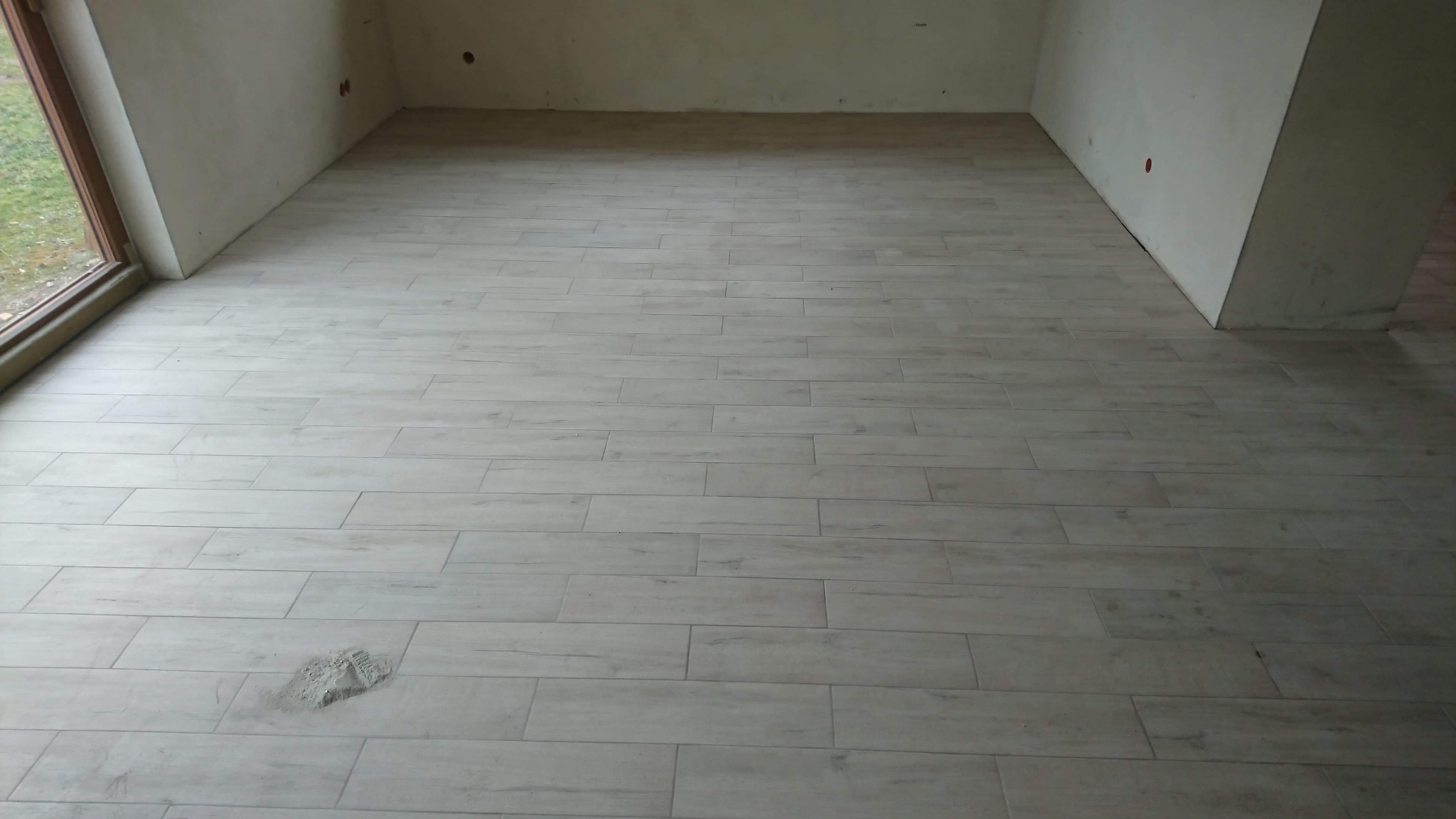 Ułożona podłoga z płytek imitujących drewno w jasnym odcieniu, widoczny fragment okna i surowych ścian, ślad zaprawy na jednej z płytek.