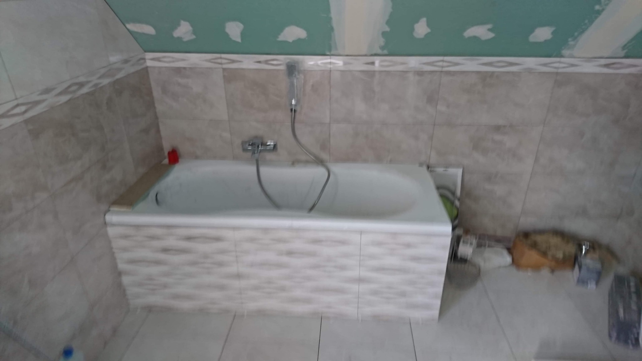 Nieukończona łazienka z wanną obudowaną płytkami, płytki na ścianach w odcieniach beżu, sufit z płyt kartonowo-gipsowych przed malowaniem, widoczne narzędzia i materiały budowlane.