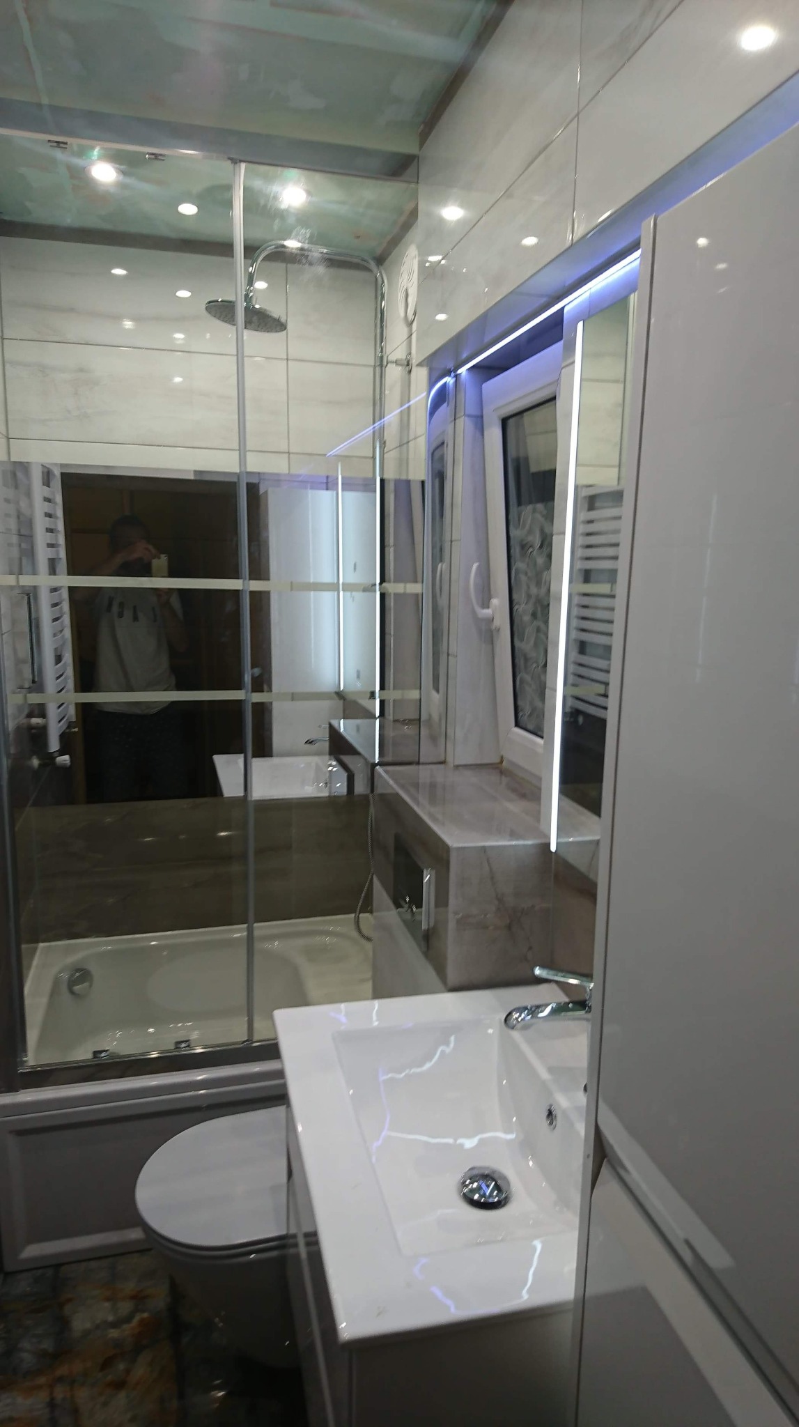 Nowoczesna łazienka z kabiną prysznicową typu walk-in, umywalką nablatową i oświetleniem LED, widoczna w lustrzanym odbiciu osoba robiąca zdjęcie.