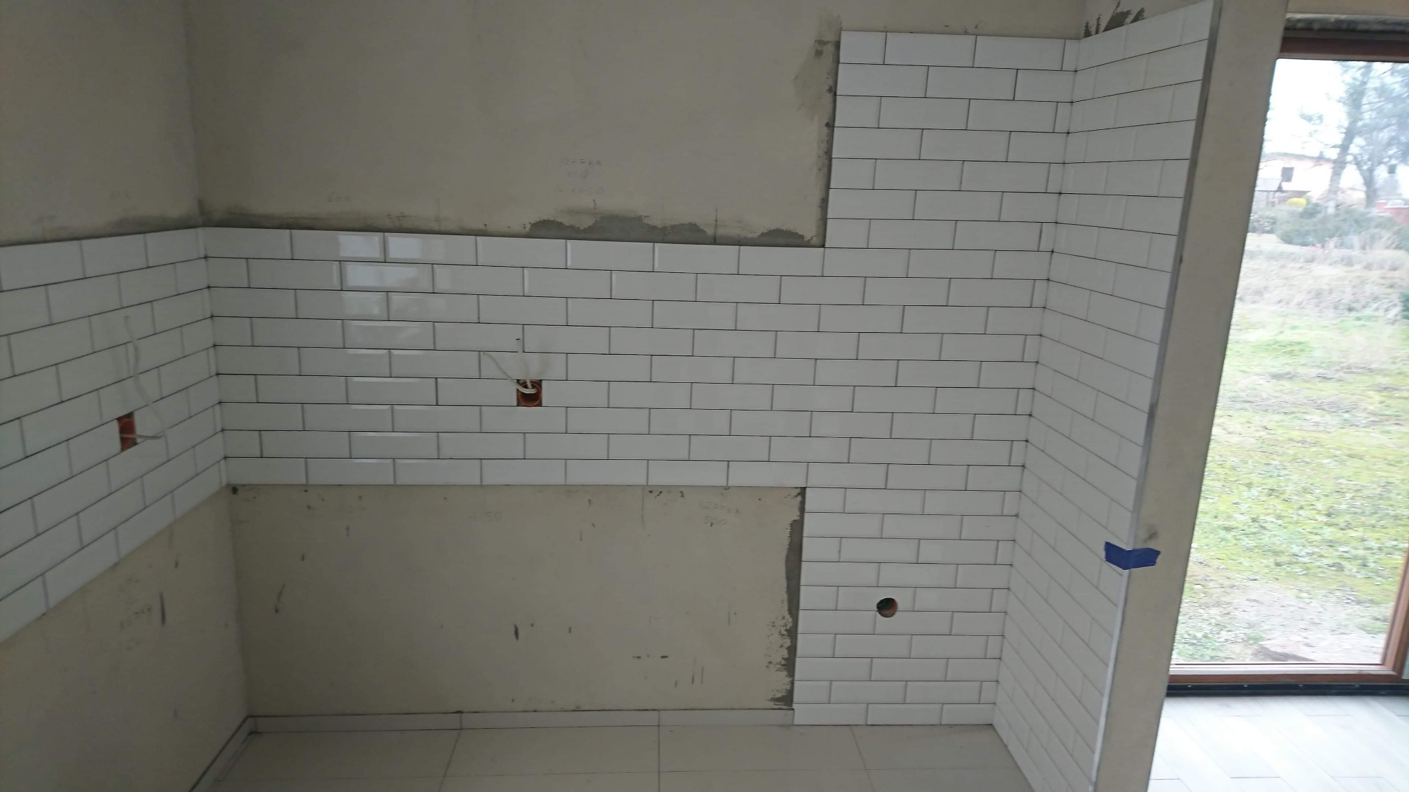 Białe, prostokątne płytki ułożone na ścianie w kuchni, częściowo jeszcze nieukończone, widoczne puszki elektryczne i otwór na rurę, okno w tle.