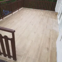 Wyłożony płytkami taras z brązową drewnianą balustradą, imitacja drewna na beżowych płytkach, widok na zielony ogród.