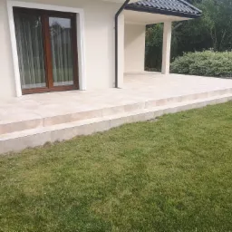 Beżowa, wykafelkowana weranda z dwoma stopniami, przylegająca do domu z brązowymi oknami i metalową barierką, otoczona trawnikiem i zielenią.