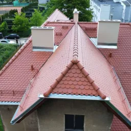 Czerwony dach z dachówki karpiówki, widok z góry. Nowe kominy, orynnowanie, zabezpieczenia przeciwśniegowe. Dach po renowacji na tle zieleni.
