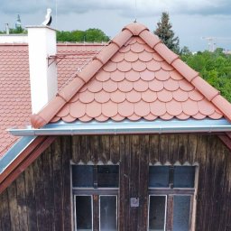 „BUD” Borucki Usługi Dekarskie Piotr Borucki - Czerwona dachówka karpiówka na drewnianym poddaszu, z bliska. Widoczne rynny, okna poddaszowe i komin z bocianem. W tle zieleń drzew i fragment panoramy miasta.