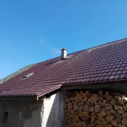 Dom z częściowo wymienionym, brązowym dachem z dachówki modułowej, widoczne przygotowanie do montażu, komin i skład drewna.