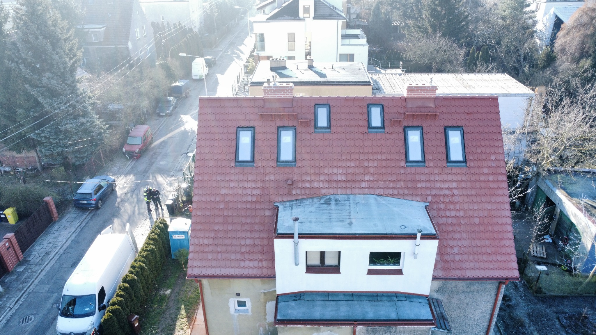 Widok z góry na dom z czerwonym dachem z dachówką i oknami dachowymi, na ulicy poniżej widoczni dwaj mężczyźni w odblaskowych ubraniach i biały van.
