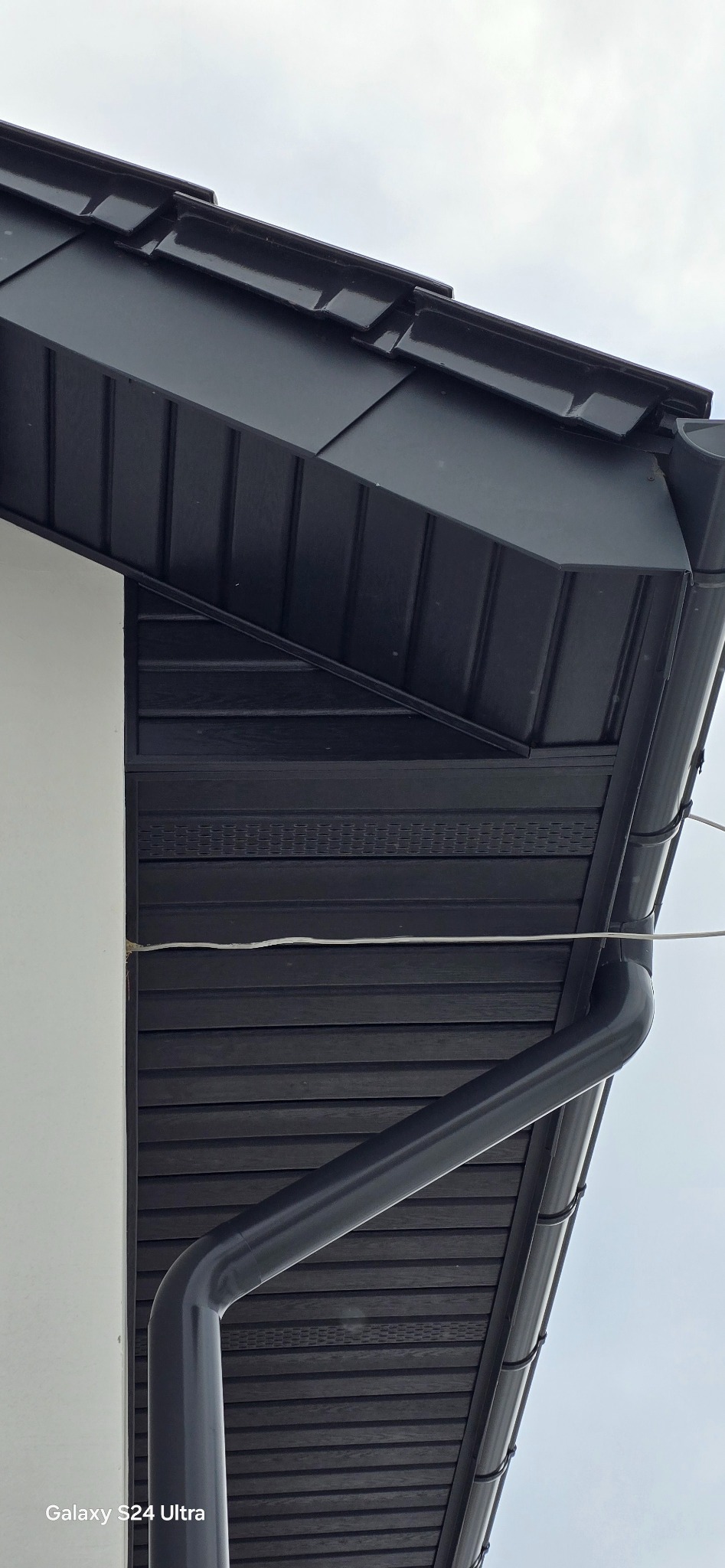 Fragment dachu z czarną blachodachówką, orynnowaniem i podbitką w kolorze grafitowym, widoczne połączenie z białą ścianą budynku.