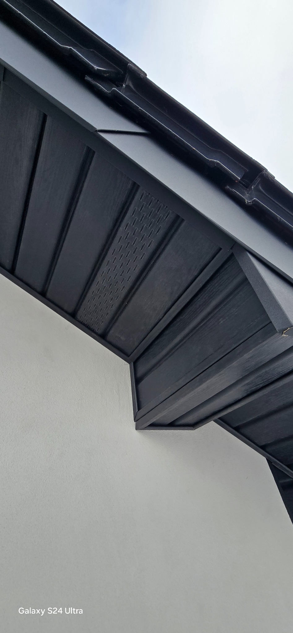 Fragment dachu z czarną podbitką z perforacją w Uciechowie, widoczne połączenie z białą ścianą budynku.