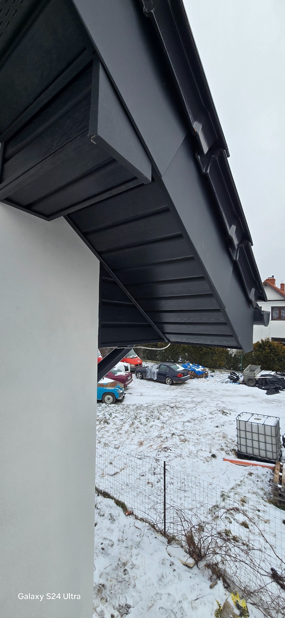 Nowe, czarne orynnowanie dachu domu jednorodzinnego, widoczne z dołu, na tle zasnieżonego podwórza z zaparkowanymi samochodami.