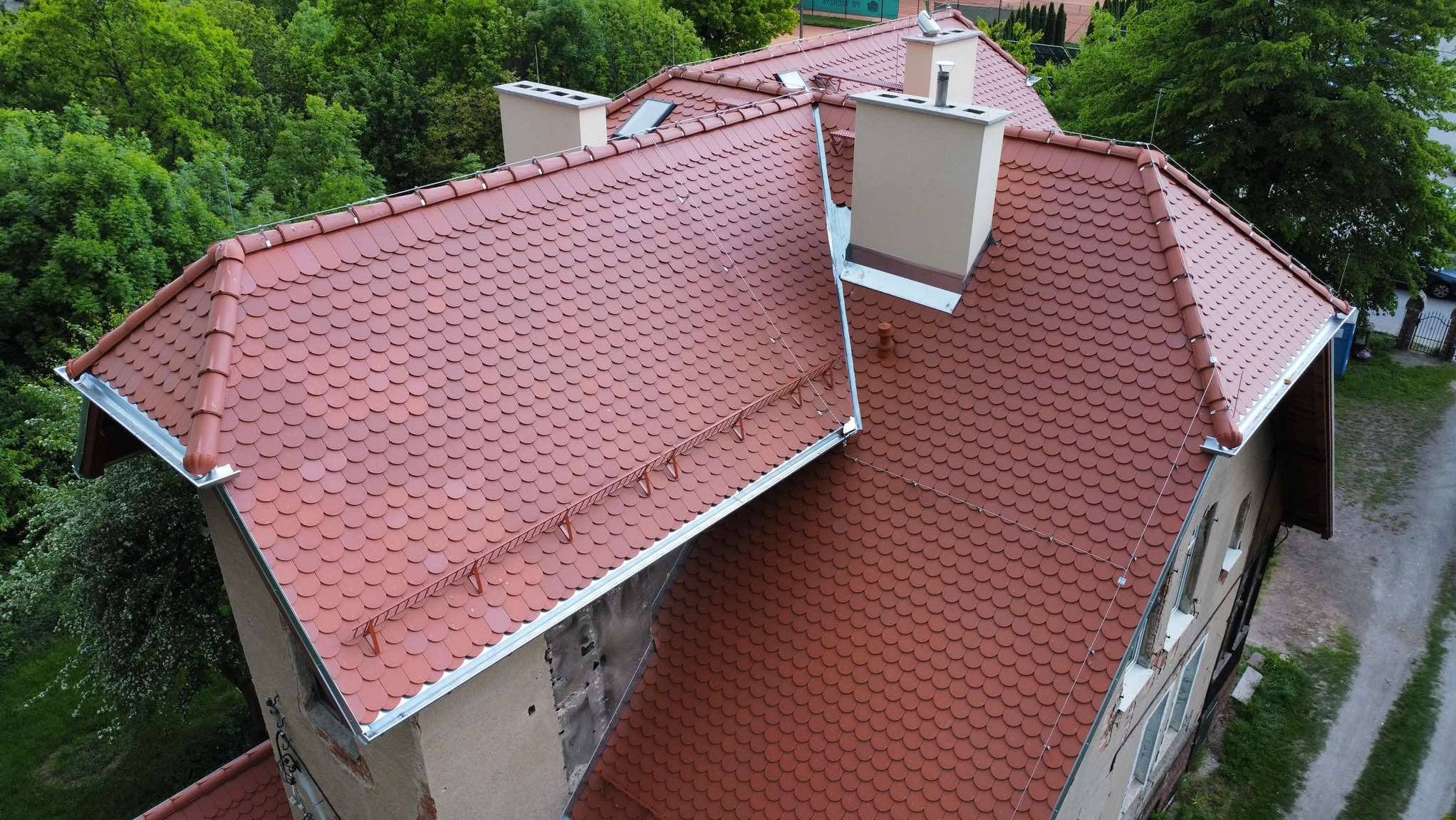 Dach pokryty czerwoną dachówką karpiówką, widoczny z góry. Nowe orynnowanie, kominy. Budynek otoczony zielenią. Charakterystyczny wzór ułożenia dachówek.