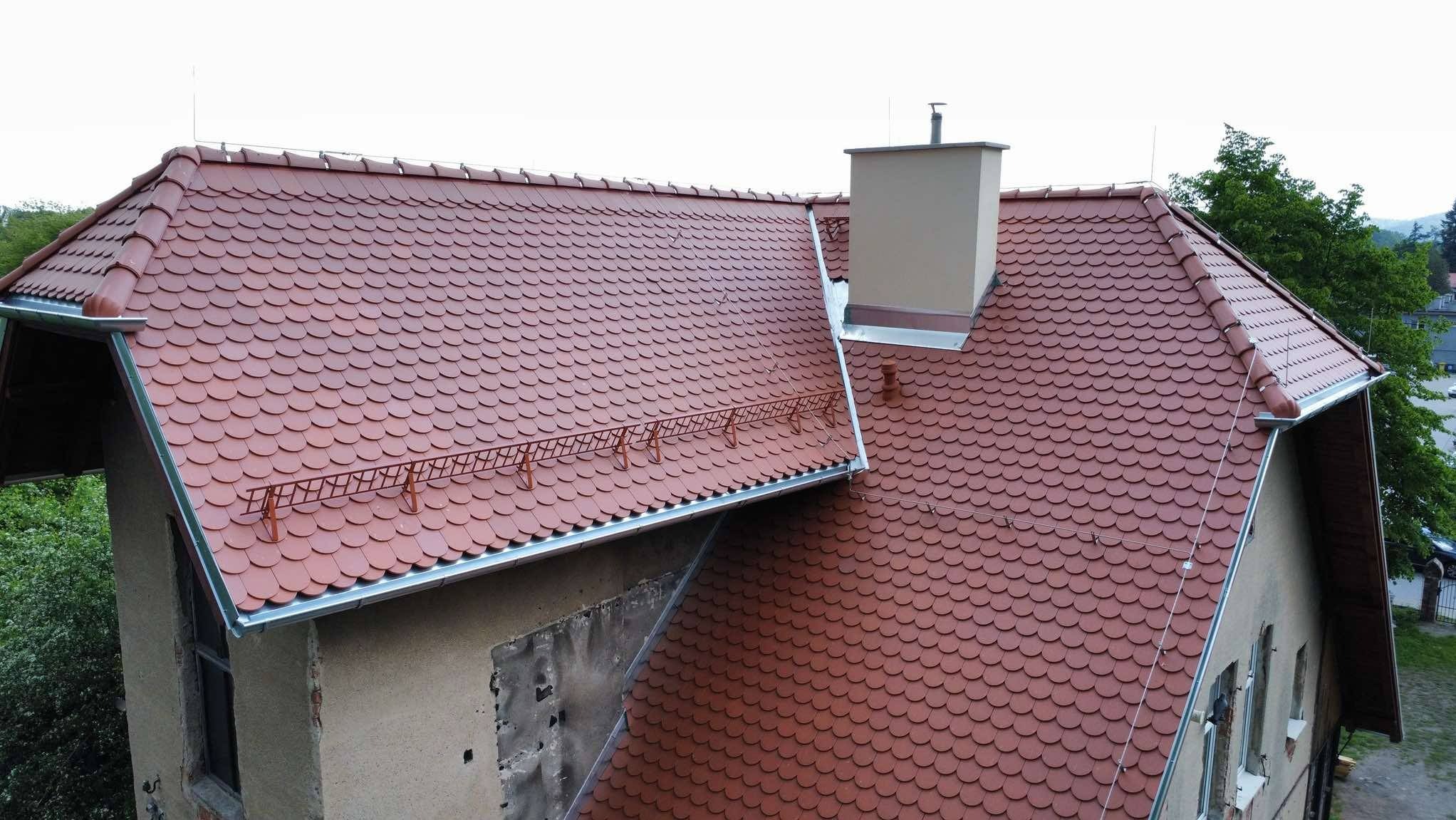 Czerwony dach z dachówki łuskowej na budynku z beżową elewacją, widoczny komin i metalowe zabezpieczenia śniegowe. Ujęcie z góry.