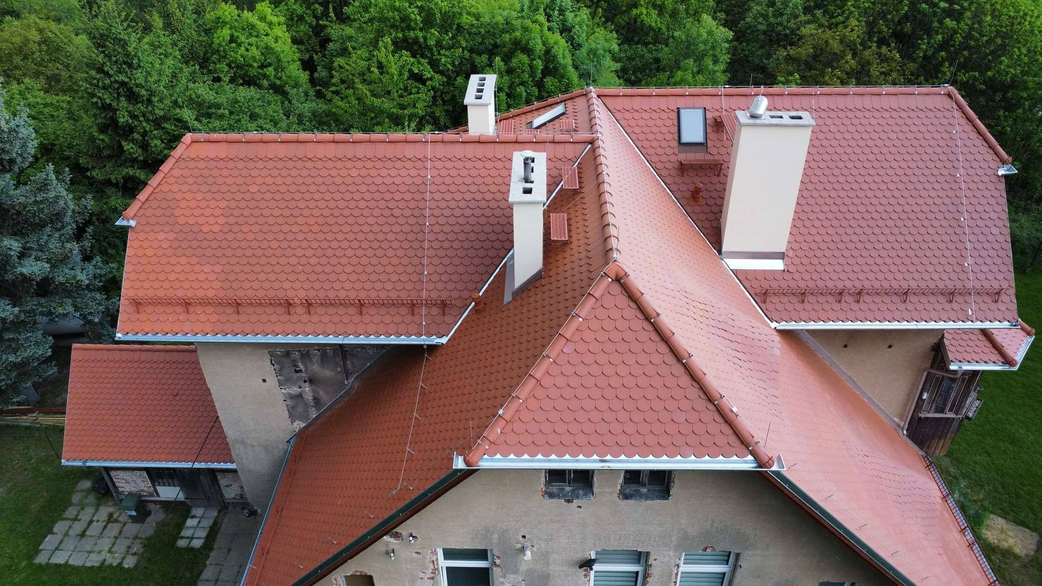 Widok z góry na budynek z nowym, czerwonym dachem pokrytym dachówką karpiówką. Widoczne kominy, okna dachowe i rynny. W tle zieleń drzew.
