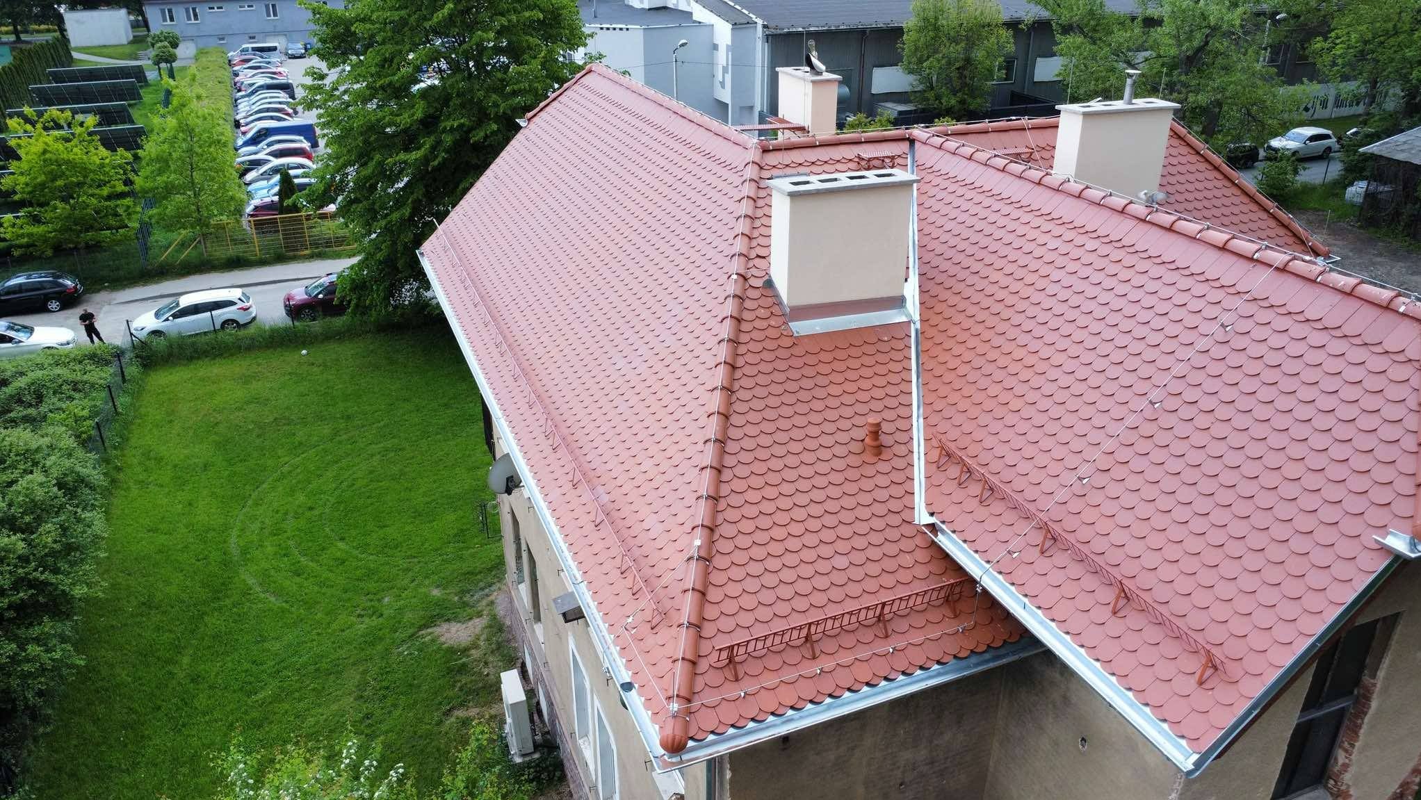 Czerwony dach z dachówki karpiówki, widok z góry na budynek z trawnikiem i parkingiem w tle. Widoczne kominy i metalowe obróbki blacharskie.