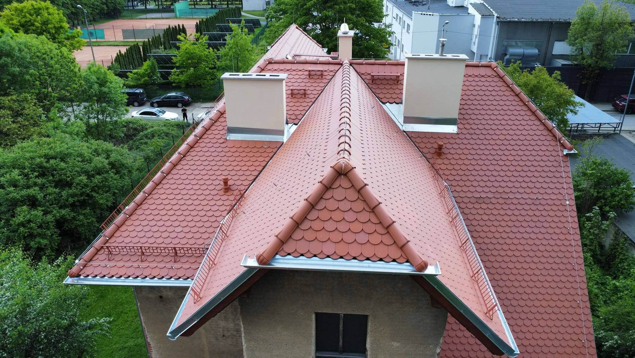 Czerwony dach z dachówki karpiówki, widok z góry. Nowe kominy, orynnowanie, zabezpieczenia przeciwśniegowe. Dach po renowacji na tle zieleni.