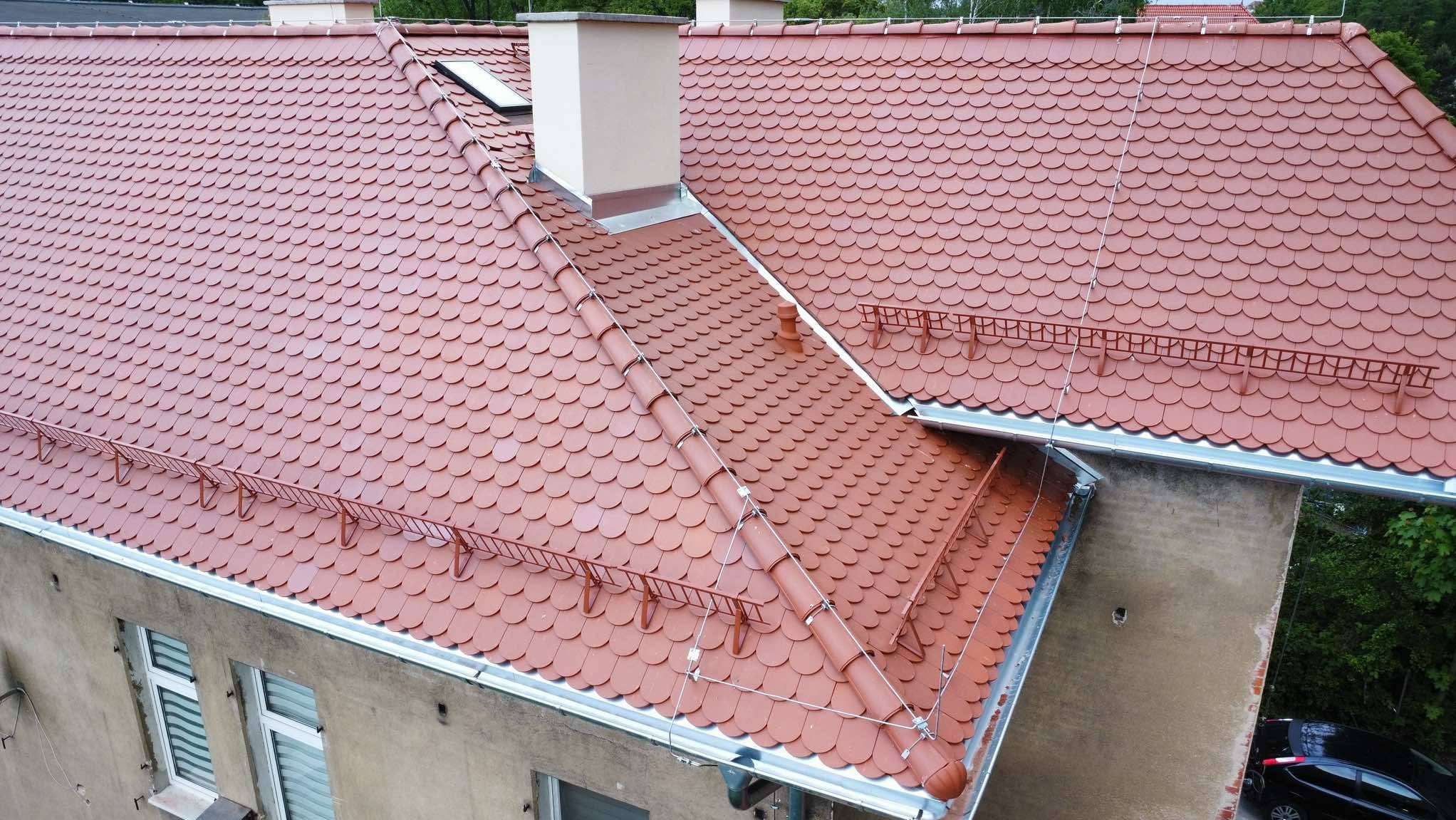 Czerwony dach z dachówki karpiówki na budynku mieszkalnym, widok z góry. Widoczne kominy, okno dachowe i system odśnieżania.