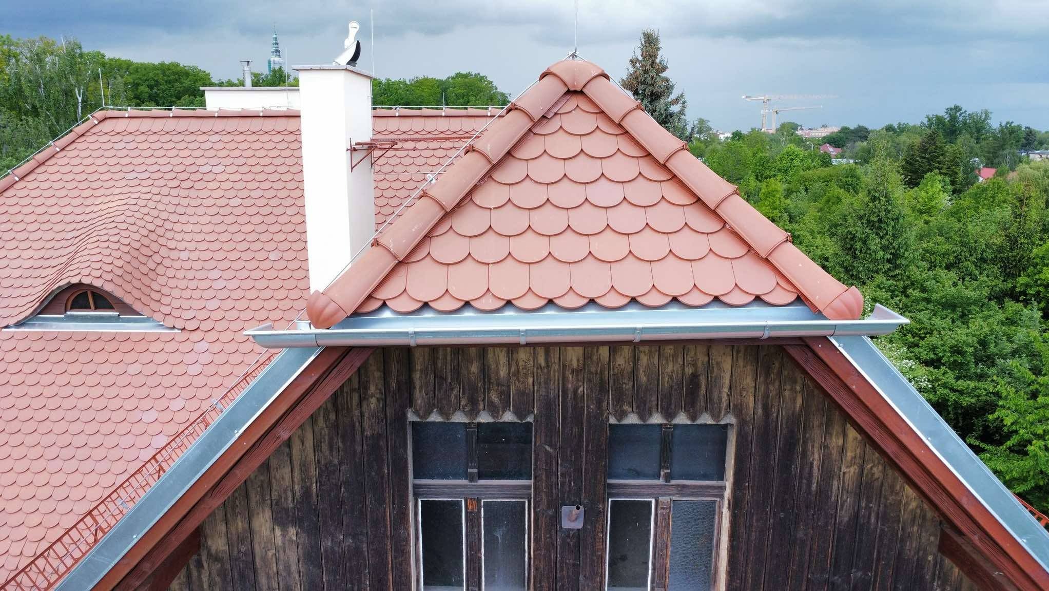 Czerwona dachówka karpiówka na drewnianym poddaszu, z bliska. Widoczne rynny, okna poddaszowe i komin z bocianem. W tle zieleń drzew i fragment panoramy miasta.