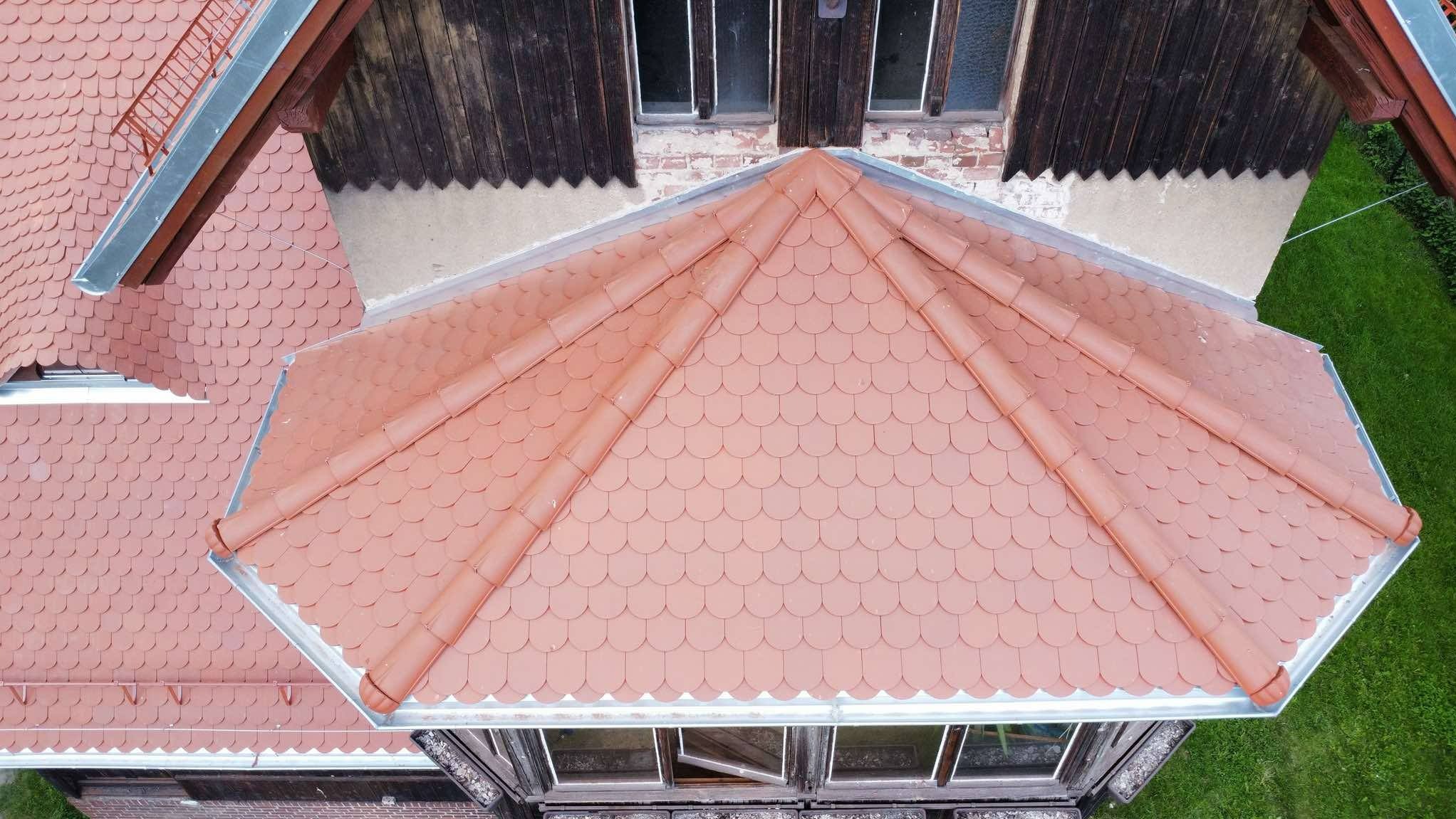 Widok z góry na fragment dachu z czerwoną dachówką w kształcie rybiej łuski i metalową rynną, z widocznymi elementami konstrukcji budynku.