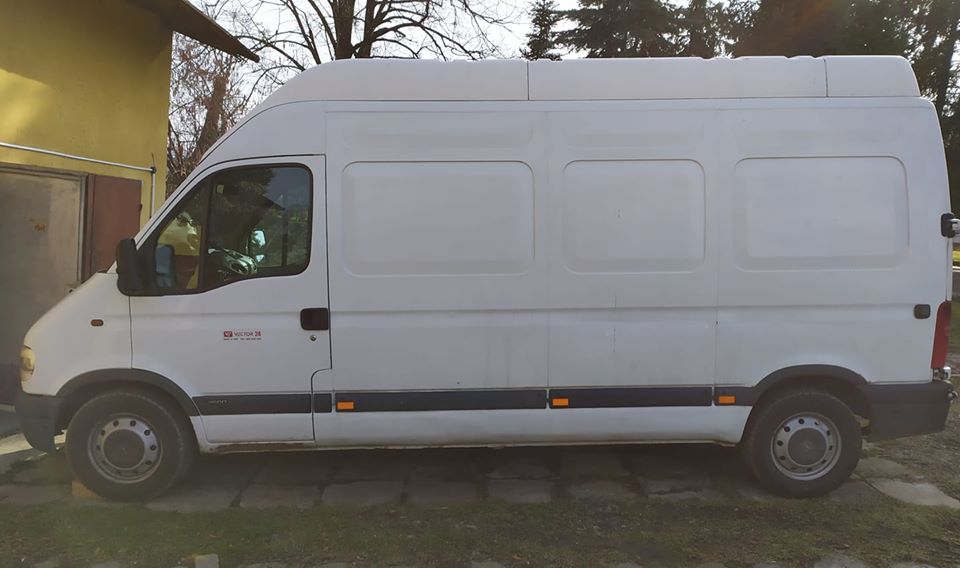 Biały, wysoki furgon dostawczy Renault Master na tle zielonego trawnika i żółtej ściany budynku gospodarczego, widok z boku.