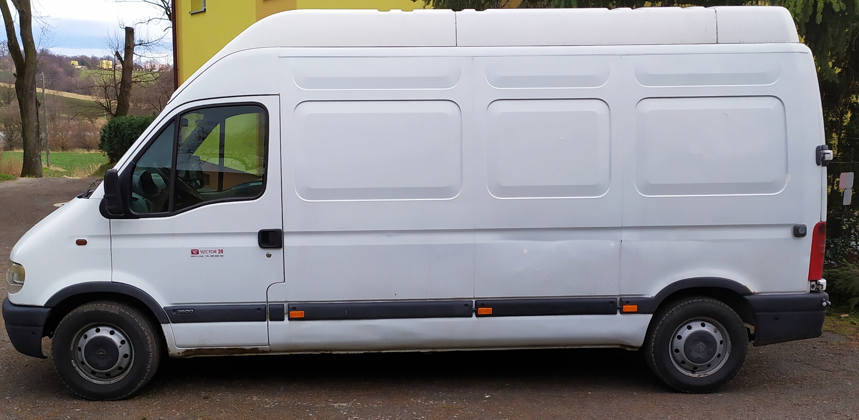 Biały samochód dostawczy Renault Master 3500 z logo Vector 24 na tle drzew i żółtego budynku.