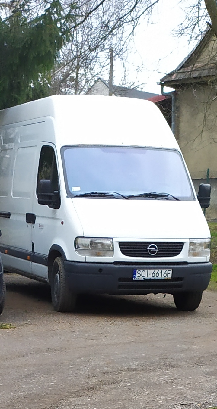 Biały Opel Movano z wysokim dachem zaparkowany na szutrowej drodze, widoczny numer rejestracyjny SCI 6616F, zieleń drzew w tle.