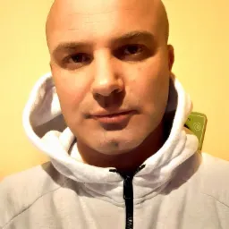 Portret mężczyzny z ogoloną głową w jasnoszarej bluzie z kapturem na tle żółtej ściany.