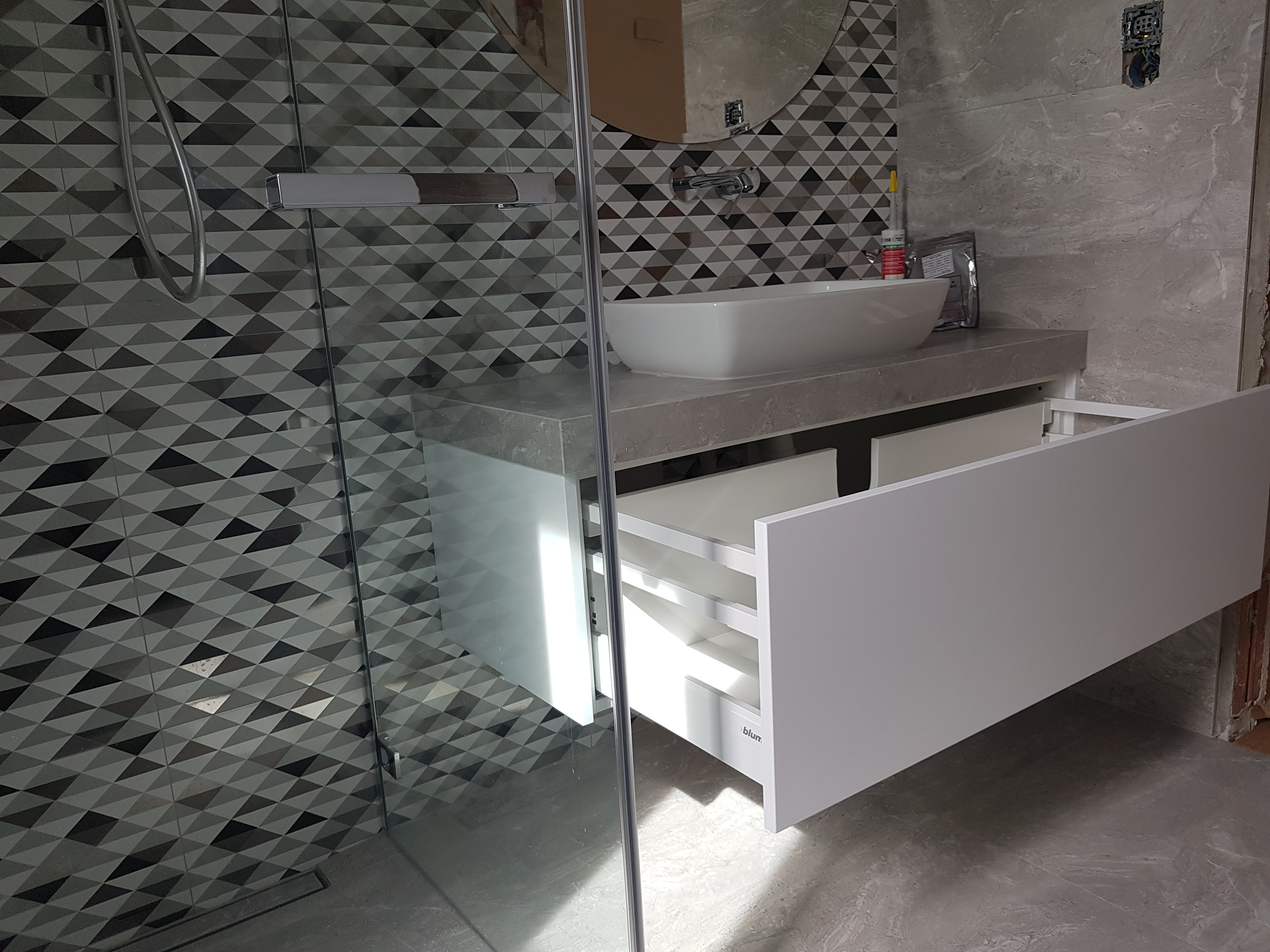 Nowoczesna łazienka w Krakowie z umywalką nablatową i białą szafką z otwartymi szufladami, widoczna przez szklaną ścianę kabiny prysznicowej z geometrycznym wzorem płytek.