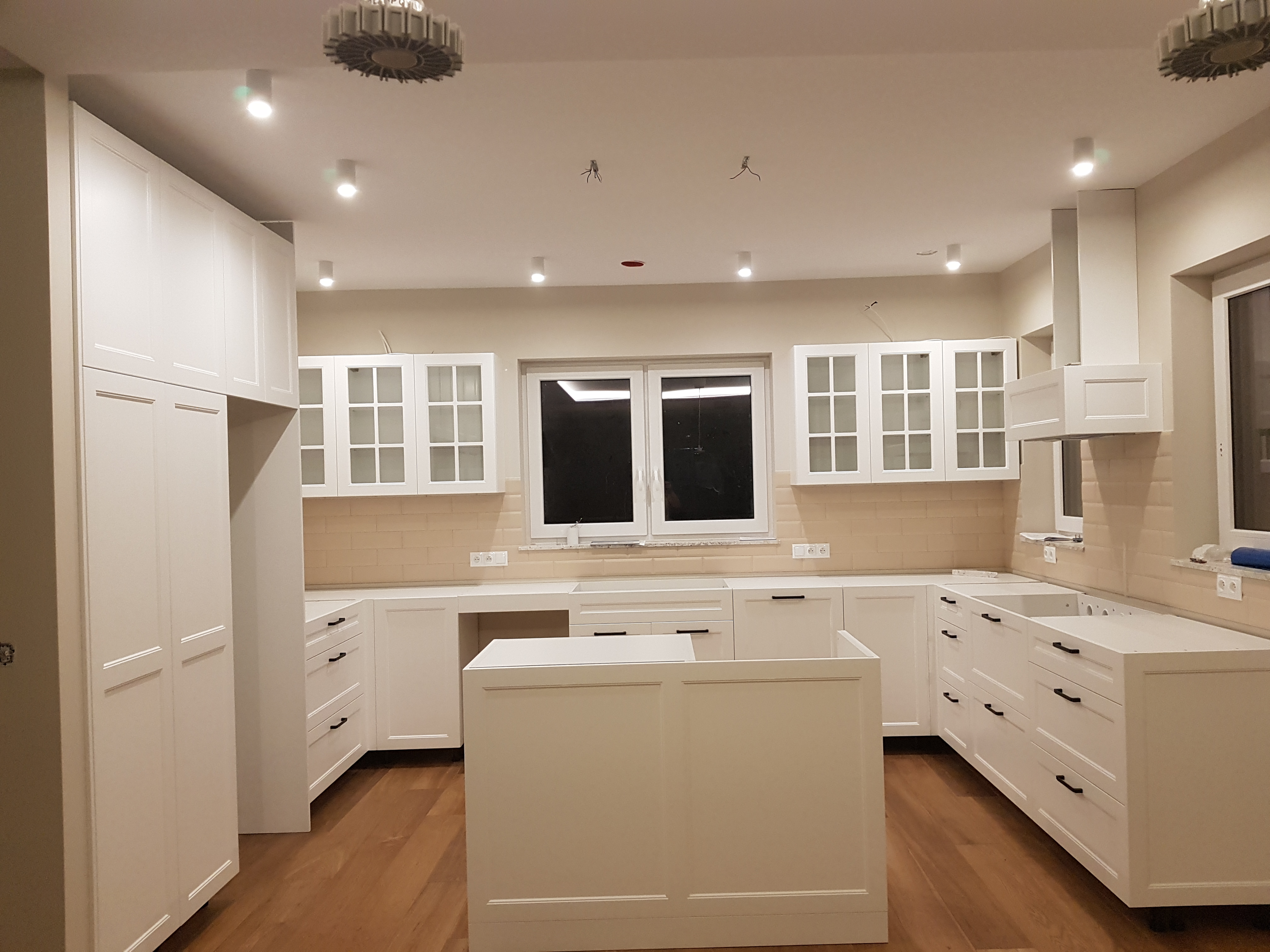 Jasna, przestronna kuchnia z białymi szafkami z frezowanymi frontami i drewnianą podłogą, wyspa kuchenna w centrum, widoczne niedokończone podłączenia elektryczne na suficie.