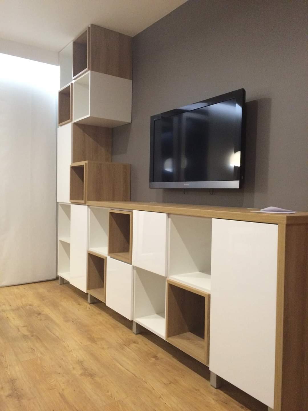Nowoczesna zabudowa ściany z szafkami i półkami w kolorze białym i dębowym, z telewizorem wiszącym na szarej ścianie w tle.
