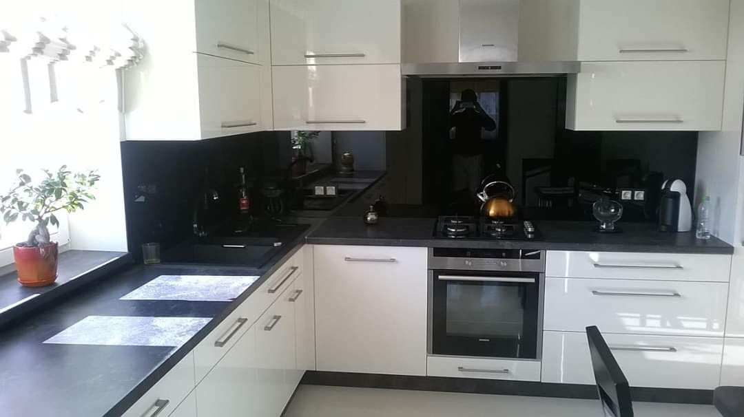 Nowoczesna kuchnia z białymi, błyszczącymi frontami szafek, czarnym blatem i szklanym panelem nad blatem, w którym odbija się osoba robiąca zdjęcie.