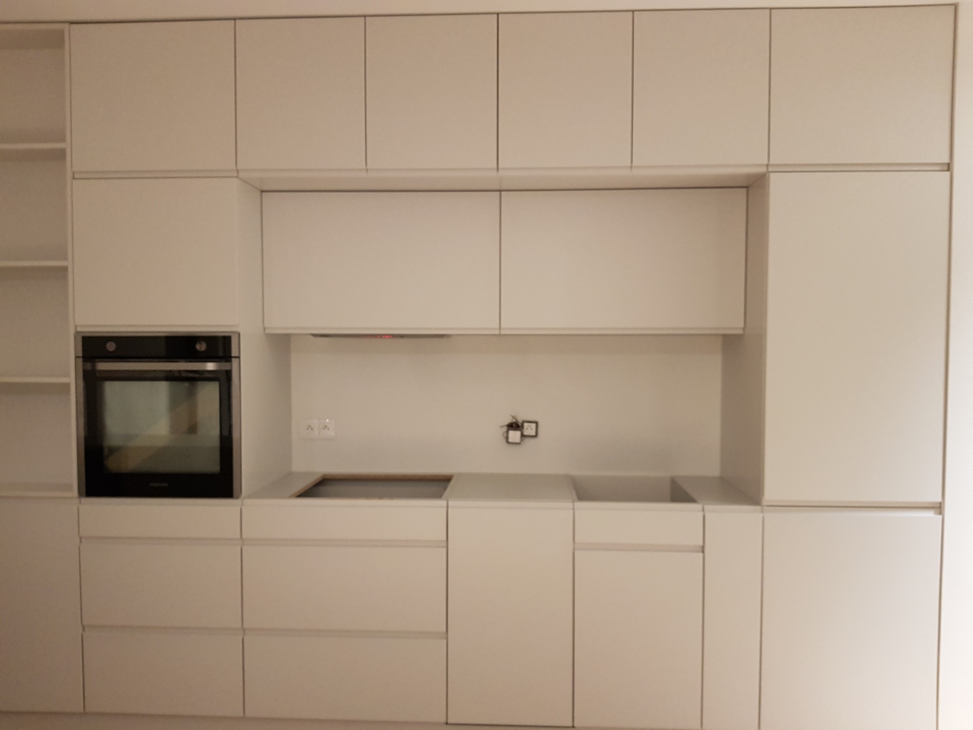 Minimalistyczna, biała zabudowa kuchenna z piekarnikiem i płytą grzewczą, bez uchwytów, z widocznymi gniazdkami elektrycznymi nad blatem.