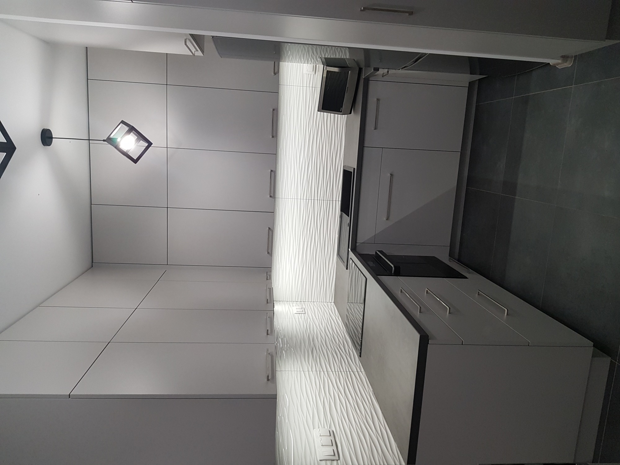 Nowoczesna, minimalistyczna kuchnia w odcieniach bieli i szarości, z frontami szafek bez uchwytów i ozdobną, teksturowaną płytką na ścianie nad blatem, oświetlona geometryczną lampą wiszącą.