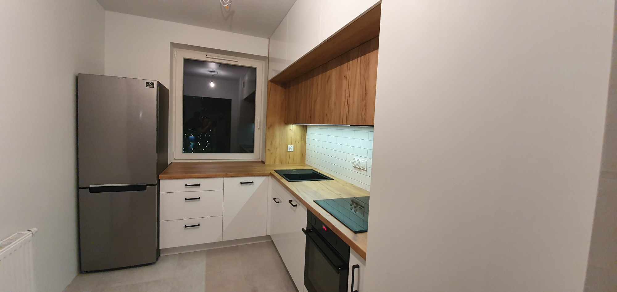 Nowoczesna kuchnia z białymi szafkami, drewnianymi blatami i szarym lodówką, widok na nocne miasto przez okno.