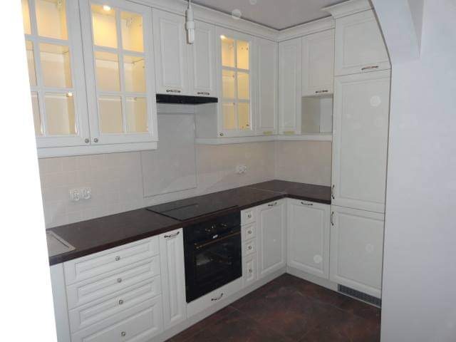 Biała, narożna zabudowa kuchenna z przeszklonymi szafkami wiszącymi i ciemnym blatem, widoczny piekarnik w zabudowie.